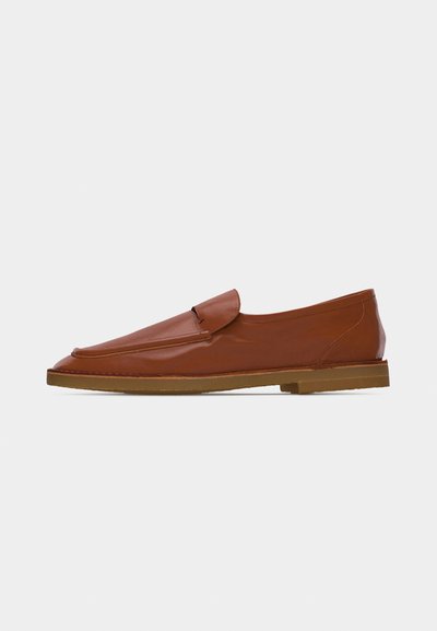 Καφέ δερμάτινο μοκασίνι slip-on με στρογγυλεμένη τετράγωνη μύτη και επίπεδη λαστιχένια σόλα, παρουσιάζεται σε απλό ανοιχτό φόντο.