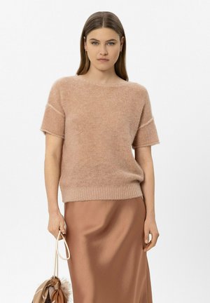 Frau trägt einen hellbraunen kurzärmeligen Strickpullover und einen passenden Satinhrock, hält eine kleine beige Handtasche mit Fellbesatz.