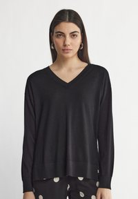 Pull noir à col V avec des manches longues, coupe ample, bords côtelés aux poignets et à l'ourlet. Texture douce, associé à un pantalon à pois noir et blanc.