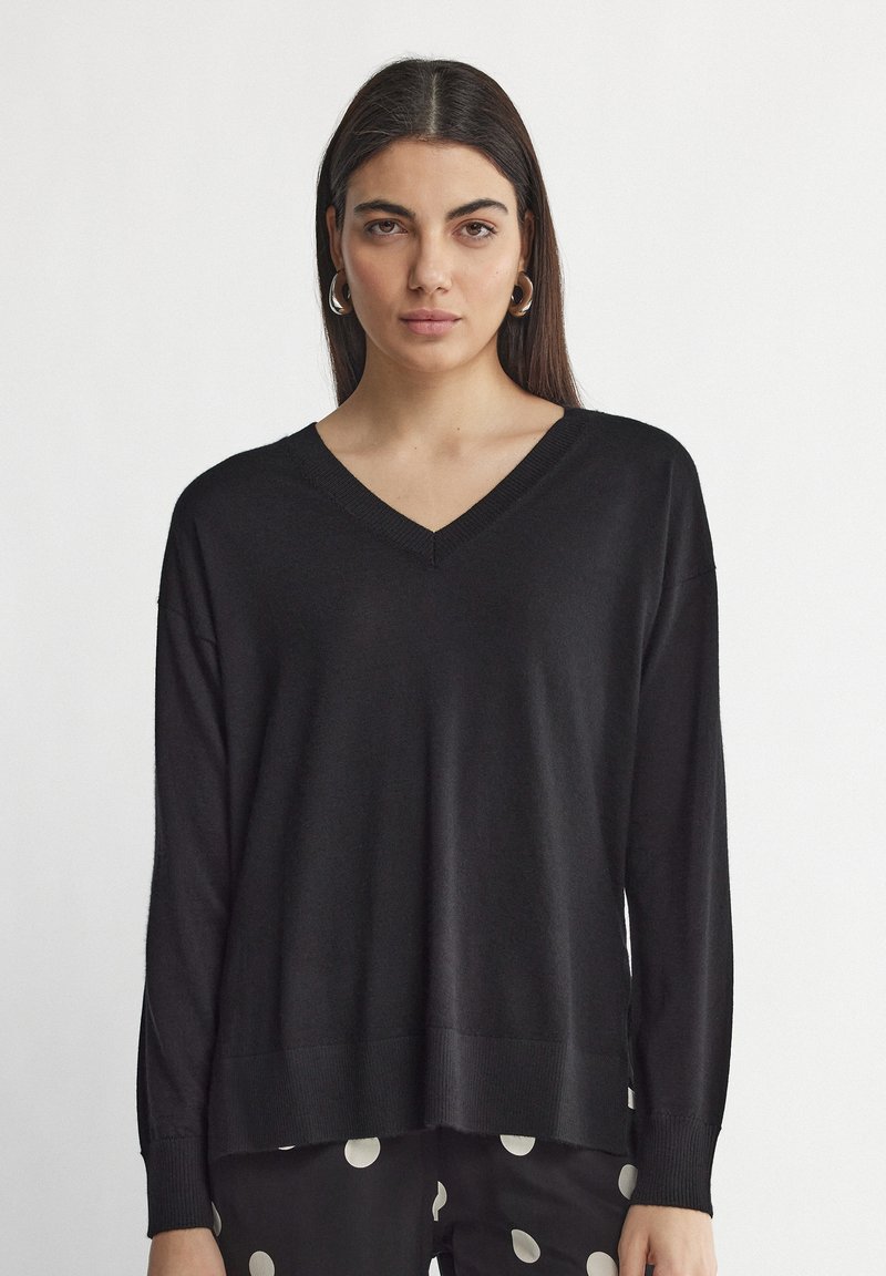 Pull noir à col V avec des manches longues, coupe ample, bords côtelés aux poignets et à l'ourlet. Texture douce, associé à un pantalon à pois noir et blanc.