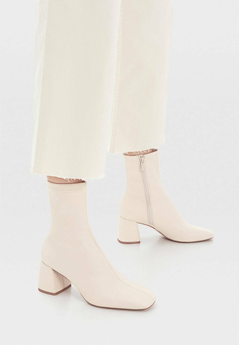 Bottes crème à hauteur de cheville avec talons carrés et fermetures éclair latérales portées avec un pantalon court écru sur un fond clair.
