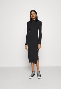 Robe en tricot côtelé noire à manches longues et fente sur le côté, portée avec des baskets montantes noir et blanc. Silhouette simple et ajustée.