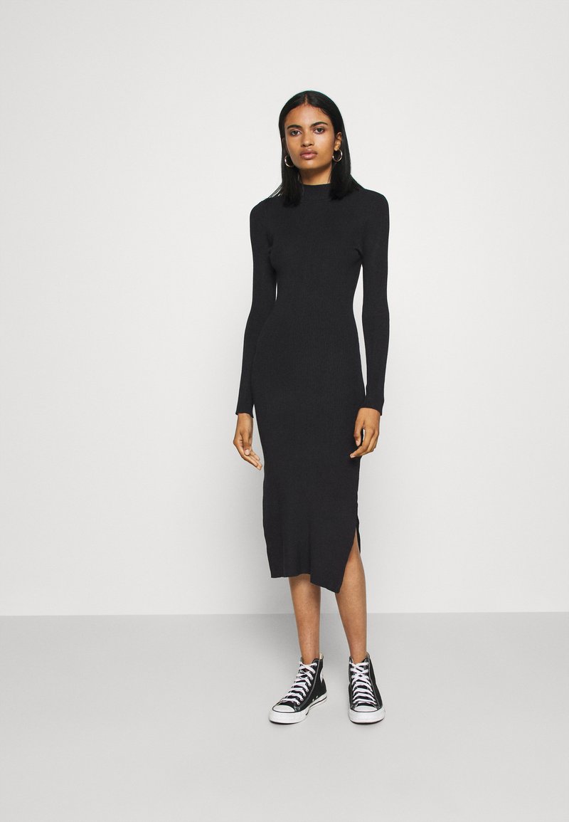 Robe en tricot côtelé noire à manches longues et fente sur le côté, portée avec des baskets montantes noir et blanc. Silhouette simple et ajustée.