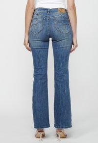 Jeans de mezclilla azules acampanados con un lavado intermedio, diseño de cinco bolsillos y bolsillos traseros bordados. Presenta un ligero desgaste y una etiqueta de cuero.