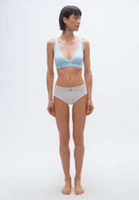 Γυναίκα που στέκεται ξυπόλητη, φορώντας ανοιχτό μπλε σουτιέν και λευκό εσώρουχο με χρυσές διακοσμητικές ζώνες στη μέση, κοιτάζοντας προς τα δεξιά, μπροστά σε λευκό φόντο.