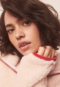 Flis jopica v nežno roza barvi z rdečim rebrastim robom; oseba s kratkimi temnimi lasmi, ki nosi jopico, roka ob bradi, prikazuje teksturo tkanine.