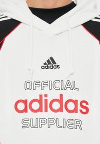 Hoodie branco com detalhes em preto e vermelho. Logótipo "adidas" em preto acima do texto "OFICIAL" e "FORNECEDOR" em letras negrito pretas e vermelhas.