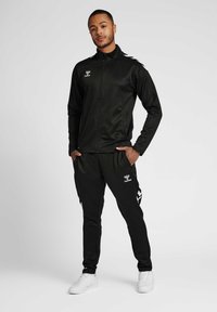 Man standing with hands in pockets, wearing black sports jacket and pants with white sneakers on plain background.