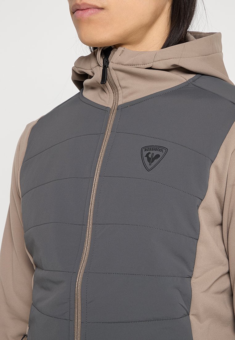 Szara i beżowa ocieplana kurtka z poziomymi przeszyciami, zamek błyskawiczny z przodu oraz logo Rossignol na klatce piersiowej. Posiada kaptur oraz miękki materiał.
