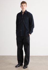 Camisa de pana azul marino con un bolsillo frontal y cierre de botones, combinada con pantalones negros y zapatos negros, sobre un fondo neutro.