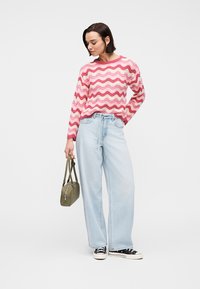 Γυναίκα που φοράει μπλούζα με ροζ και λευκές κυματιστές ρίγες, ανοιχτόχρωμα μπλε τζιν παντελόνια με φαρδιές μπατζάκια, μαύρα αθλητικά παπούτσια και κρατά μια μικρή τσάντα σε ελιά πράσινο χρώμα.