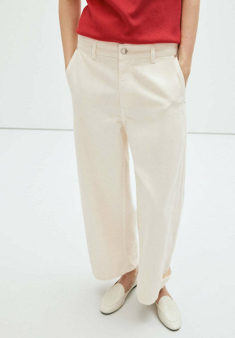 Personne portant un pantalon crème à jambes larges, des chaussures blanches type slip-on et un haut rouge, les mains dans les poches, sur fond blanc.