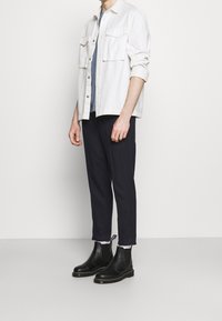 Chemise blanche boutonnée avec deux poches poitrine, portée sur un t-shirt bleu, associée à un pantalon bleu marine foncé et des bottines Chelsea noires.