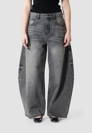Personne portant un jean en denim noir délavé à jambes larges et des chaussures plates noires ornées de perles, debout devant un fond clair uni.