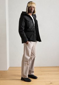 Parka negra con capucha de piel sintética, cierres de cremallera y botones a presión, cuatro bolsillos frontales; combinada con pantalones beige claros y zapatos negros.