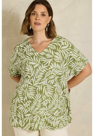 Femme portant un haut à col en V avec un motif de feuilles vert et blanc, manches courtes, et un pantalon beige sur un fond neutre.
