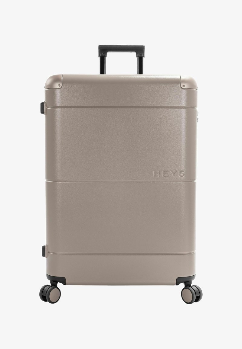 Valise rigide beige avec quatre roues multidirectionnelles et poignée noire rétractable, nom de la marque "Heys" embossé sur le devant.