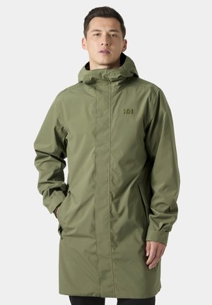 Uomo che indossa una lunga giacca da pioggia verde oliva con cappuccio, zip frontale e mani nelle tasche laterali, in piedi davanti a uno sfondo uniforme.
