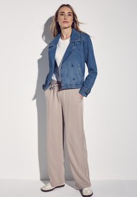 Ljust blå denimjacka med dubbelknäppning, vit t-shirt under, beiga vida byxor och vita sneakers. Slät textur.