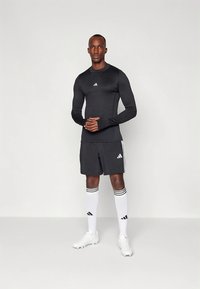 Fekete hosszú ujjú sportfelső, fekete rövidnadrág és fehér zokni fekete csíkokkal; az Adidas logói láthatók a ruházaton és a cipőkön.