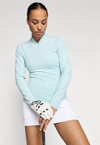 T-shirt azul claro de manga longa com um design de meia zíper, textura suave. Combinado com uma saia branca, usando uma luva de golfe branca, segurando um taco.