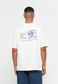 Man in een wit T-shirt met paarse graphic van een fluitje dat rennend is, houdt een sjaal vast met de tekst "HSW" en de tekst "Kick Off Club, Serving the Game for Generations".