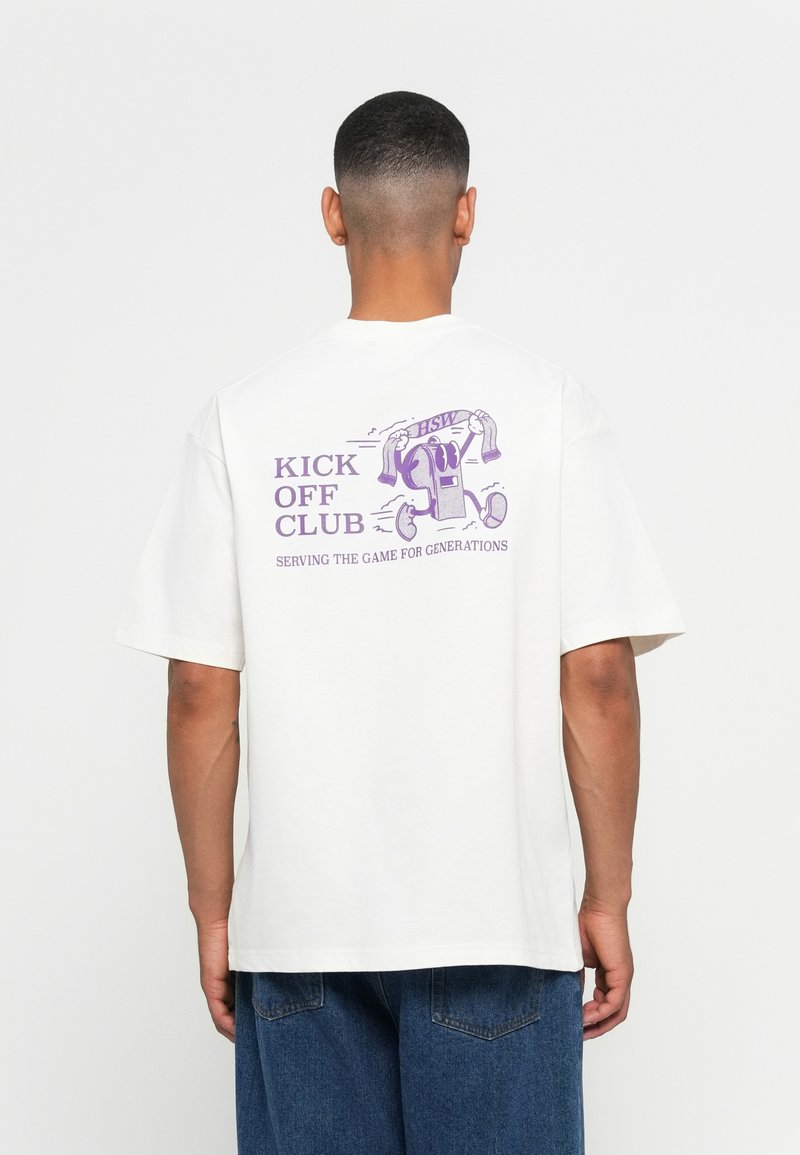 Man in een wit T-shirt met paarse graphic van een fluitje dat rennend is, houdt een sjaal vast met de tekst "HSW" en de tekst "Kick Off Club, Serving the Game for Generations".