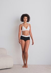 Bijela bralette i crni donji dio s visokim strukom; glatka tkanina; jednostavan, minimalan dizajn; model stoji nasuprot jednostavnom zidu.