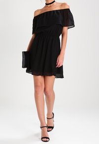 Schwarzes schulterfreies Kleid aus schichtweisem Chiffonstoff, elastischer Taille und ausgestelltem Saum. Kombiniert mit schwarzen Absätzen und einer schwarzen Clutch.