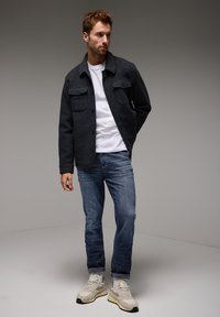 Mörkgrå ulljacka med två bröstfickor, vit T-shirt med rund halsringning, blå jeans och ljusa sneakers mot en neutral bakgrund.