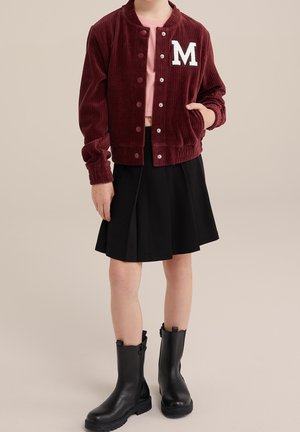 Kind draagt een bordeauxrode corduroy jas met een "M", een roze cropped shirt, een zwarte geplooide rok en zwarte laarzen tot halverwege de kuit, staat met een hand in de zak.