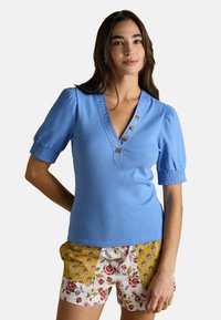 Sieviete, tērpusies zilā īsām piedurknēm virsajā un ziedu raksta šortos, stāv ar vienu roku aiz muguras.