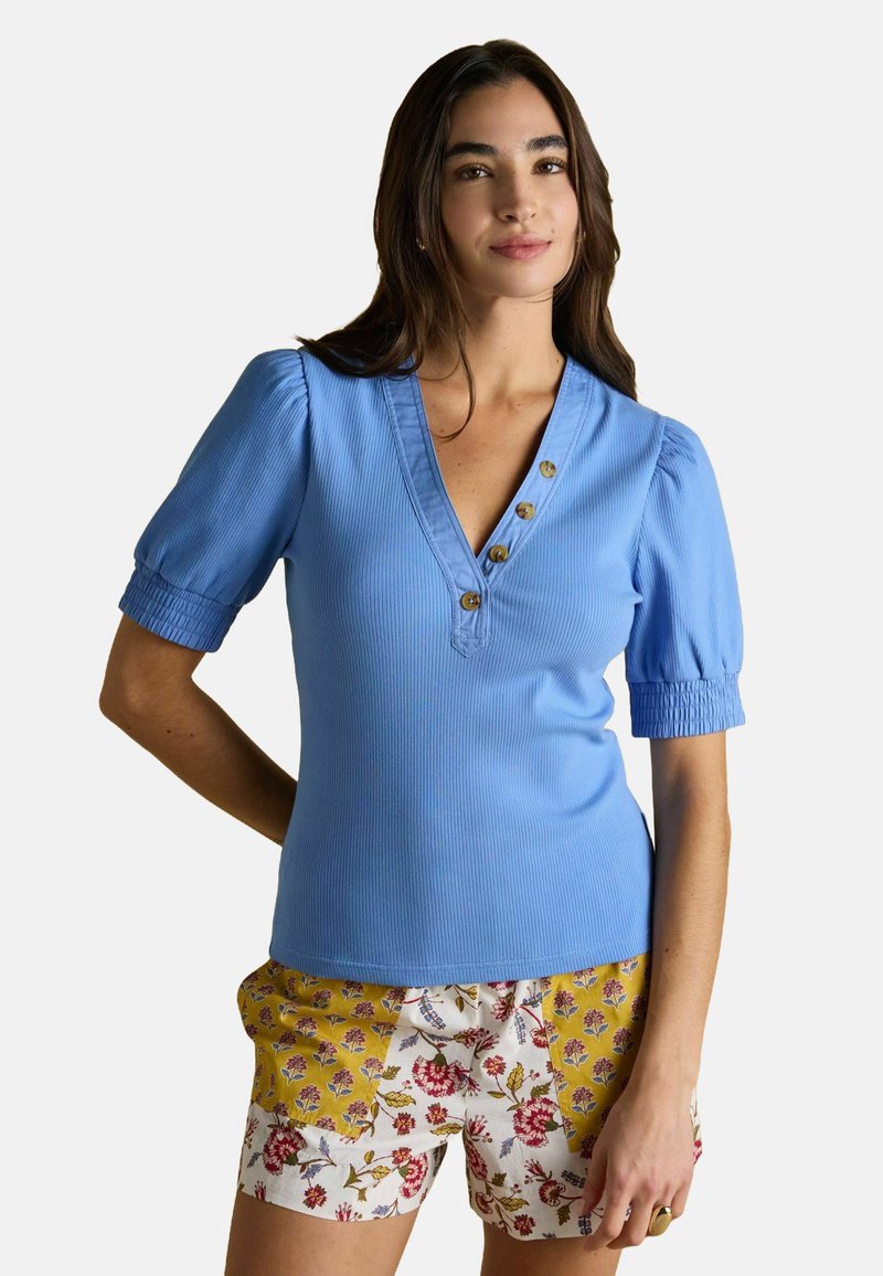 Sieviete, tērpusies zilā īsām piedurknēm virsajā un ziedu raksta šortos, stāv ar vienu roku aiz muguras.