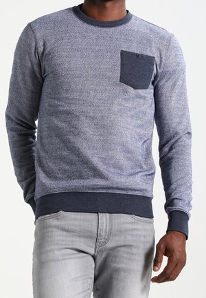 Mann trägt einen blauen gemusterten Pullover mit langen Ärmeln, einer dunklen Brusttasche und hellgrauen Jeans, steht mit einer sichtbaren Hand.