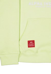Felpa verde chiaro con tasca e etichetta rossa Alpha Industries, con testo parziale bianco "Alpha Ind Knoxville, Tennessee" sopra.
