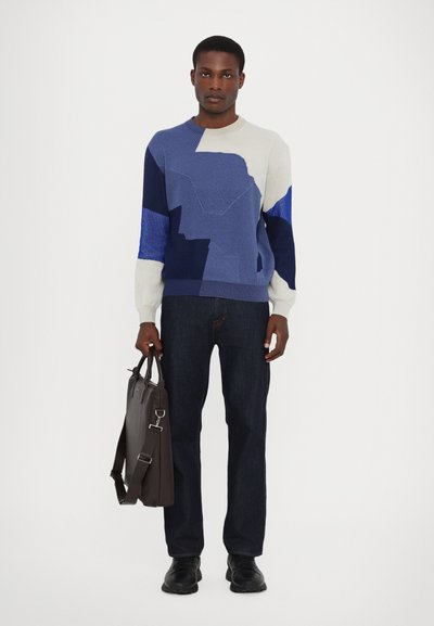 Knit sweater in blue, cream, and navy with geometric patterns; paired with dark jeans and holding a brown leather briefcase.