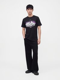 Jeune homme debout, mains dans les poches, portant un T-shirt noir à motif graphique, un pantalon noir à jambes larges et des baskets noires sur fond blanc uni.