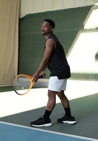 Teniso žaidėjas su juoda be Rankovių marškinėliais, baltais šortais ir juodais sportbačiais, laikantis oranžinį teniso raketę ant korto.