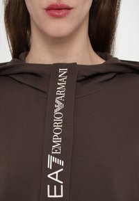 Brūns kapucēs krekls ar kapuci, kam priekšpusē ir izteikta balta logo etiķete ar tekstu "EA7 Emporio Armani" serif burtu stilā.