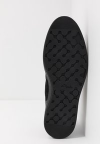 Czarna gumowa podeszwa buta z teksturowanym, falistym wzorem i zaokrąglonymi kształtami. Widoczne logo po stronie, zaprojektowana z myślą o przyczepności i trwałości.