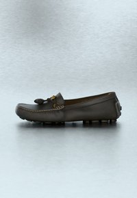 Mocassin en cuir noir avec des détails cousus, un pompon sur le dessus et des semelles en caoutchouc avec grip, présenté de profil sur fond gris.