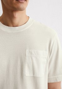 Jasnobeżowy t-shirt z krótkim rękawem wykonany z teksturowanej bawełny, z okrągłym dekoltem i jedną naszywaną kieszenią na lewej piersi.