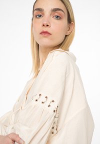 Blusa beige chiaro con un tessuto morbido e testurizzato, caratterizzata da maniche arricciate, dettagli in occhio di pernice e accentuazioni in metallo argentato.