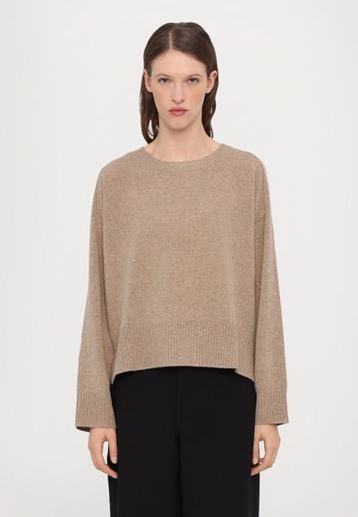 Lysebrune strikket trøje med rund halsudskæring, dropped shoulders og ribbede manchetter. Har en cropped pasform og blød struktur. Sorte bukser synlige.