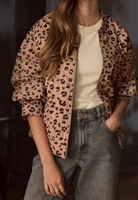 Kurtka bomber z nadrukiem w cętki lamparta w jasnoróżowym materiale, z zamkiem z przodu i oversize’owymi rękawami, zestawiona z beżowym prążkowanym topem i wysokim stanem dżinsami.