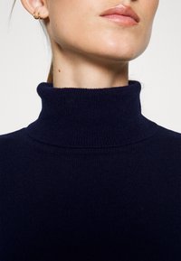 Tmavě modrý rolák z ribovaného úpletu. Má vysoký, složený límec a minimalistický design bez viditelných vzorů nebo akcentů.