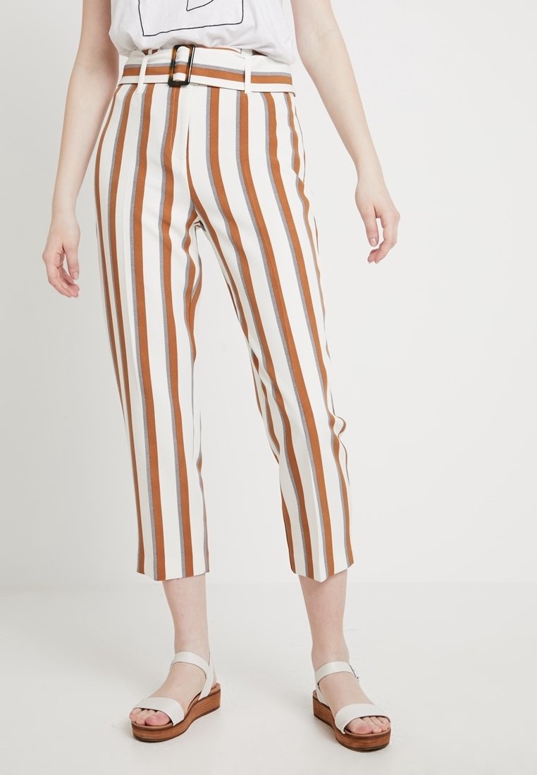 Femme portant un pantalon court taille haute avec des rayures verticales blanches, brunes et grises, assorti à des sandales blanches à semelles en bois.
