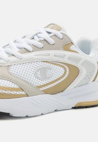 Scarpa sportiva bianca con pannelli in suede e rete beige. Presenta una suola testurizzata, perforazioni e un logo sul lato. I lacci sono bianchi.