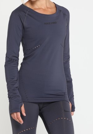 Woman wearing dark gray Nike Pro long-sleeve top and matching leggings with perforated detail and thumbholes, standing against a plain background.