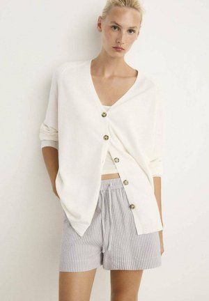 Cárdigan blanco de canalé con escote en V y cinco botones frontales. Combinado con pantalones cortos a rayas en gris y blanco, con cinturilla elástica.